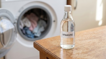 Botella de vidrio con vinagre sobre una mesa de madera. Detrás, una lavadora de ropa blanca con la puerta abierta que contiene prendas de colores.