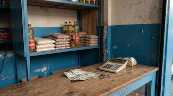 Mostrador de madera con billetes de dólar y monedas, una calculadora antigua, y estantes azules con bolsas de arroz, frijoles y botellas de aceite.