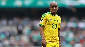 Hombre calvo, tez oscura, con camiseta y pantalón corto de fútbol amarillo y verde con el número 23. Fondo de estadio borroso