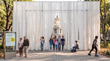 Una estructura con cortinas blancas en un parque público, por donde caminan varias personas con el Palacio de Bellas Artes al fondo.