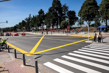 Intersección de asfalto con un paso de peatones de franjas blancas, demarcación amarilla, bolardos y dos personas en la acera