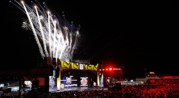 Un escenario iluminado durante la noche con fuegos artificiales en el aire y una multitud de espectadores en el público