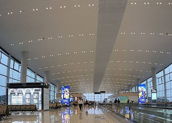 Interior de una terminal de aeropuerto grande y moderna con techo blanco, luces empotradas, grandes ventanales y pantallas digitales. Personas caminan y esperan
