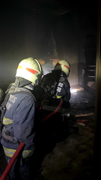 Dos bomberos con cascos y trajes protectores avanzan en un espacio oscuro y humeante con escombros quemados, apuntando una manguera