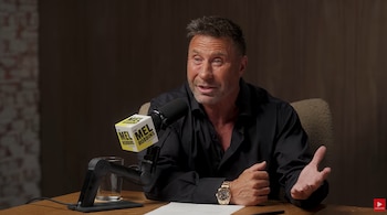 Un hombre con camisa negra y reloj dorado habla frente a un micrófono en una mesa de estudio