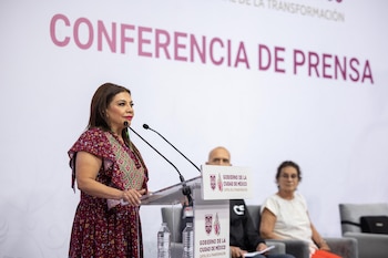 Mujer con vestido rojo floral habla en un podio transparente con dos micrófonos, frente a una pared blanca con el texto 'CONFERENCIA DE PRENSA'