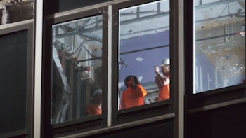 Ventanas de un edificio. Dentro, varios trabajadores de construcción con cascos blancos y chalecos naranjas. Estructuras metálicas expuestas