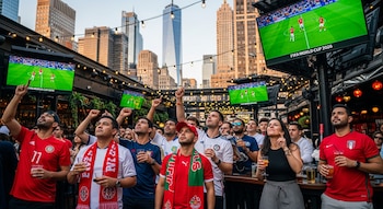 Un grupo de personas en un bar al aire libre en Nueva York observa pantallas con un partido de fútbol y el texto "FIFA World Cup 2026". Edificios altos al fondo.