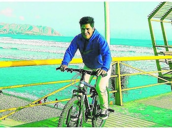 Un hombre con sudadera azul y pantalón claro conduce una bicicleta en un malecón junto al mar y colinas al fondo