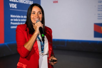 Una mujer de cabello oscuro y blazer rojo sonríe mientras sostiene un micrófono y un control remoto. Detrás hay una pantalla de proyección con texto en azul y blanco