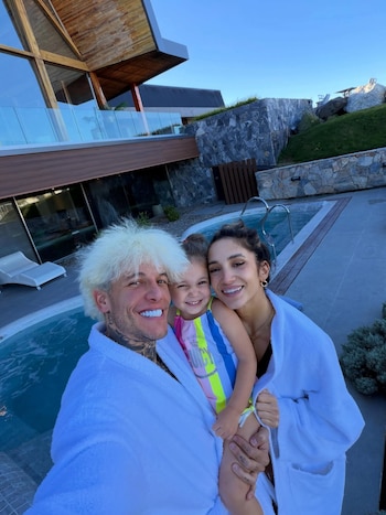 Alex Caniggia, Melody Luz y su hija Venezia posando sonrientes con batas blancas junto a una piscina al aire libre, con una casa moderna de fondo