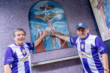 Dos hombres con camisetas de fútbol moradas y blancas tocan un mosaico religioso de Jesucristo en una pared con azulejos morados