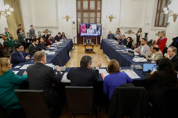 Marinos Kartikkis y Jorge Quiroz, vestidos de traje, se dan la mano y sonríen a la cámara en una sala grande con decoraciones doradas y medios de comunicación