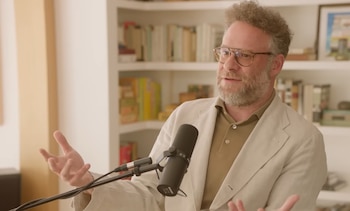 Seth Rogen con gafas y barba, lleva una chaqueta beige y polo marrón, sentado frente a un micrófono, gesticulando. Un estante con libros al fondo