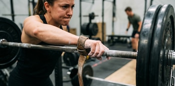Primer plano de una mujer con correas de cuero en las muñecas, sujetando una barra con discos de pesas en un gimnasio