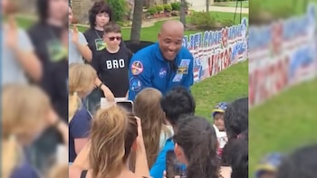 Un hombre calvo sonriente con un traje de astronauta azul de la NASA está rodeado de niños que sostienen teléfonos, con decoraciones de banderas de EE. UU. en el fondo
