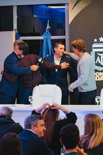 Cuatro hombres en un escenario; dos abrazándose, uno sonriendo, otro de espaldas. Mesa, sillas, copas con agua, público y pantalla con logo AFA