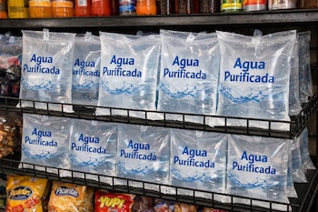 Varias bolsas transparentes de agua purificada con letras azules, llenas de líquido, dispuestas en dos estantes metálicos negros en una tienda