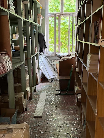 Interior de una biblioteca dañada. Fragmentos de vidrio en el suelo, estanterías de madera con libros y cajas, y un trozo de material de construcción. Ventana rota