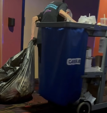 Persona mayor de espaldas sacando una bolsa de basura negra grande junto a un carro de limpieza azul en un pasillo de cine con paredes rosas