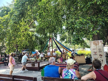 Personas en un evento al aire libre; grupo de músicos en escenario de madera con telas colgantes; puestos de mercado al fondo, bajo grandes árboles