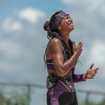 Mujer atleta con el rostro y cuerpo sucios, vestida con ropa deportiva morada, levanta las manos y mira hacia arriba con una expresión de intensa emoción bajo el cielo.
