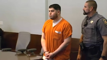 Un hombre esposado con uniforme naranja permanece de pie en una sala judicial junto a un agente policial uniformado