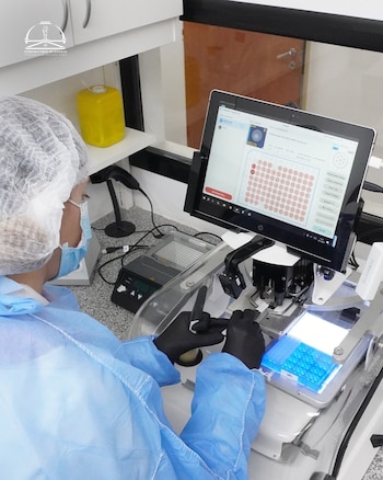 Una persona con gorro, mascarilla y bata azul, guantes negros, opera un equipo de laboratorio con una pantalla que muestra una cuadrícula de círculos