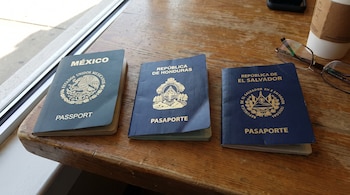 Tres pasaportes cerrados, uno verde y dos azules, sobre una mesa de madera. Se observan gafas y un vaso de café en el lateral derecho.