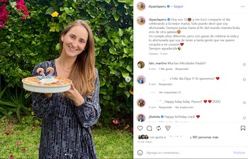 Mujer joven con cabello castaño y blusa oscura sonríe, sostiene un postre en un molde blanco con velas azules y rosadas con el número 32, en un jardín con plantas verdes y flores