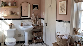 Un baño con bañera, inodoro, lavabo de pedestal, espejo, estantes de madera, cestas de mimbre, paredes rosas y suelo de mosaico blanco y negro.