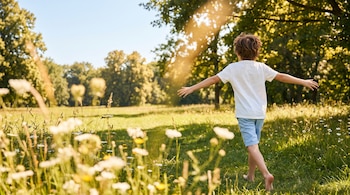 Niño de espaldas corriendo descalzo en un parque soleado sobre césped verde. Viste remera blanca y short azul. Flores silvestres desenfocadas en primer plano.