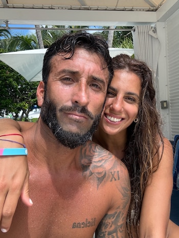 Un hombre con barba y tatuajes posa para una fotografía junto a una mujer sonriente de largo cabello castaño al aire libre