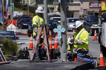 Tres trabajadores con casco y chaleco amarillo realizan obras junto a una escalera y un poste metálico con una cruz azul pegada