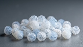 Primer plano de un montón de microesferas blancas y azul claro con textura porosa, apoyadas sobre una superficie lisa gris con fondo desenfocado