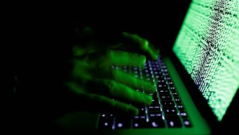 Primer plano de manos escribiendo en un teclado de portátil, con la pantalla iluminada por código verde en un entorno oscuro