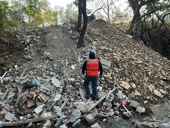 Hombre de espaldas con chaleco naranja y gorro azul inspecciona un gran vertedero de escombros y basura a la orilla de un pequeño arroyo en un entorno natural