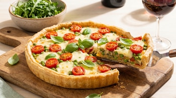 Vista aérea de una tarta salada con tomates cherry, muzzarella fundida y arvejas sobre una tabla de madera, mientras se retira una porción.