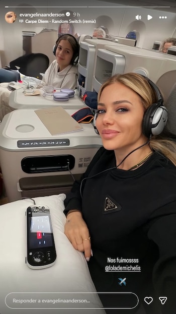 Una mujer rubia y una niña sentadas en asientos de avión de primera clase, ambas llevan auriculares. Se observa un control remoto de entretenimiento