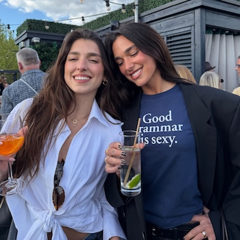 Dua Lipa, con una camiseta azul y blazer negro, sonríe junto a una mujer con camisa blanca y bebida naranja en un patio al aire libre