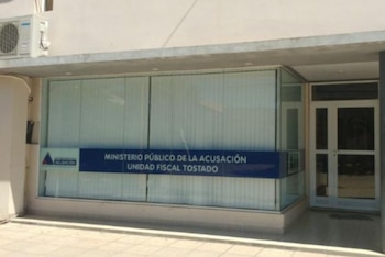 Exterior de un edificio con ventanas, cartel azul 'Ministerio Público de la Acusación Unidad Fiscal Tostado', aire acondicionado y puerta blanca