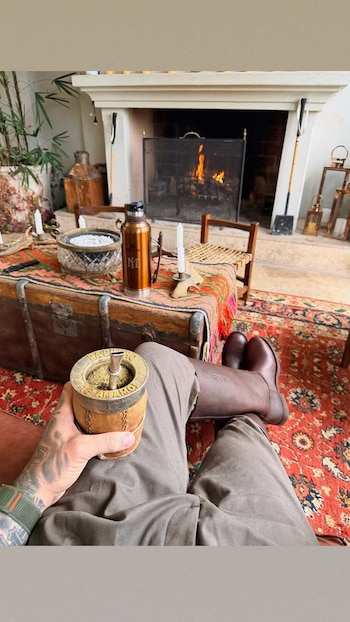 Una mano tatuada sostiene un mate grabado frente a una mesa con un termo y una chimenea encendida al fondo