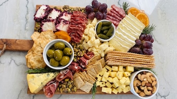 Tabla de madera con quesos variados, fiambres, uvas rojas, naranjas, pistachos, anacardos, almendras, aceitunas, pepinillos, galletas y romero.