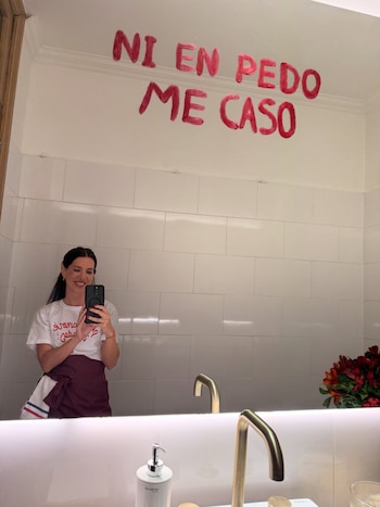 Una mujer toma una foto con un teléfono móvil frente a un espejo que tiene la frase «NI EN PEDO ME CASO» escrita en letra roja