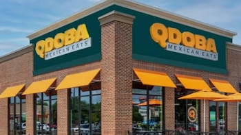 Un edificio de ladrillos con ventanales grandes, toldos naranjas y letreros amarillos de Qdoba Mexican Eats. Hay mesas, sillas y sombrillas afuera