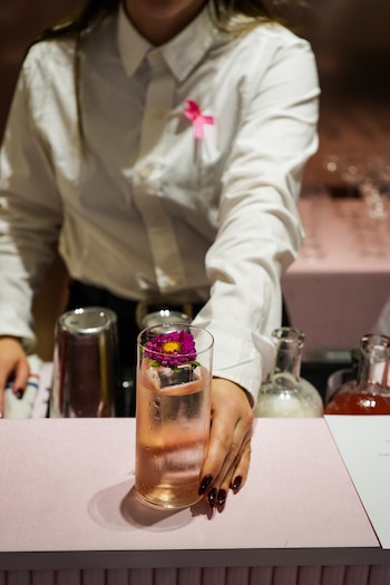 Persona con camisa blanca y lazo rosa sirve un vaso alto con una bebida rosada, un hielo y una flor en la barra