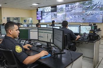 Sala de control con varios operadores uniformados frente a múltiples monitores de computadora. Una gran pared de video muestra imágenes en vivo de cámaras de tráfico urbano.