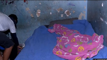 Una cama con cobija azul y manta rosa junto a paredes de pintura deteriorada, mientras una persona agachada revisa el suelo