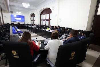 Tres personas sentadas ante una mesa con micrófonos, carpetas con documentos y un letrero de identificación en una sala oficial