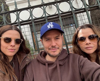 Tres personas posan frente a una reja metálica; dos mujeres llevan gafas de sol y el hombre del centro usa una gorra azul